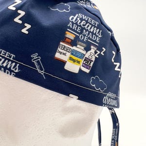 Op de afbeelding: Een blauwe scrub-muts met een witte print van flesjes met de labels "VERSED", "PROPOFOL" en "PDEX" met de tekst "Sweet dreams are made of..." en "zzz" die overal herhaald wordt.