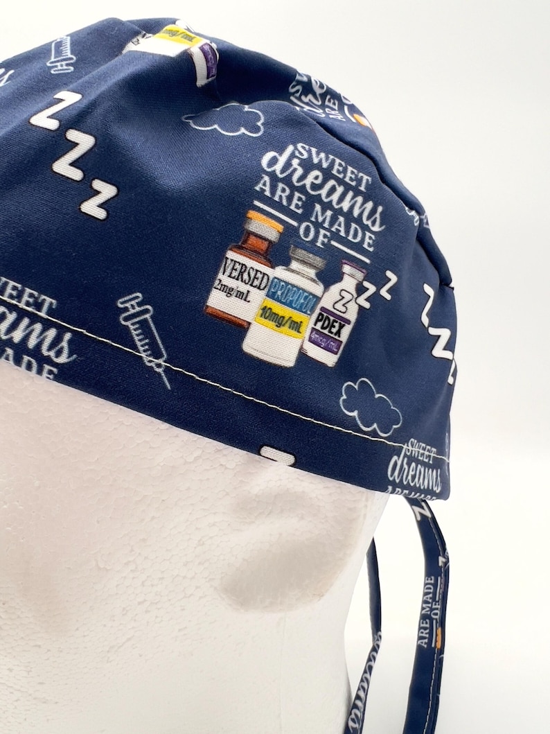 Op de afbeelding: Een blauwe scrub-muts met een witte print van flesjes met de labels "VERSED", "PROPOFOL" en "PDEX" met de tekst "Sweet dreams are made of..." en "zzz" die overal herhaald wordt.