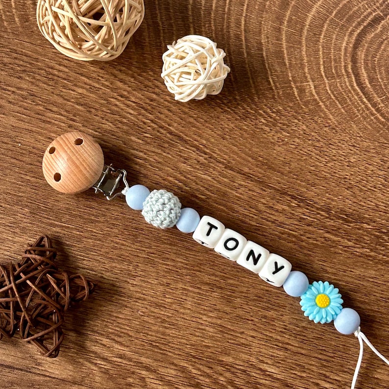 May include: A blue and white baby teether with the name "TONY" spelled out in white letters on blue beads. The teether has a wooden clip and a blue flower bead.