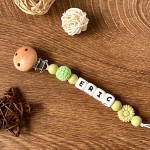 May include: A wooden clip with a green and white silicone beaded chain with the name "ERIC" spelled out in white beads. The chain has a green crocheted bead and a sunflower charm.