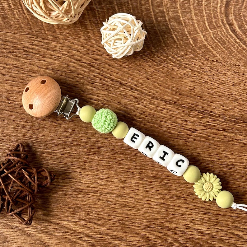 May include: A wooden clip with a green and white silicone beaded chain with the name "ERIC" spelled out in white beads. The chain has a green crocheted bead and a sunflower charm.