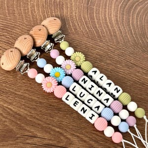 Puede incluir: Cuatro clips de chupete personalizados con cierres de madera y metal. Cada clip presenta una cadena de cuentas de colores, incluyendo blanco, rosa, azul y verde, con acentos florales. Los nombres "LENI", "LUCA", "NINA" y "ALAN" están escritos con cuentas de letras blancas.