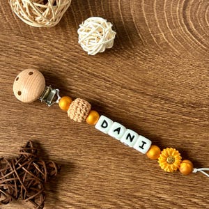 May include: A wooden baby teether with a clip, beads, and a crocheted ball. The beads are gold and white and spell out the name "DANI". There is a yellow sunflower charm on the teether.