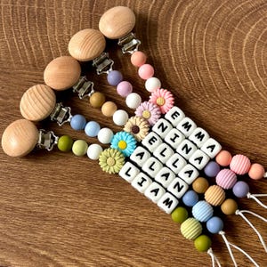 Puede incluir: Cinco mordedores para bebés de silicona y madera de colores con clips. Cada mordedor tiene un nombre diferente escrito en letras blancas: Emma, Nina, Ella, Alan y Liam. Los mordedores están decorados con pequeños amuletos de flores.