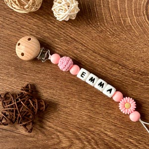 May include: Pink and white baby teether with the name "EMMA" spelled out in white beads. The teether has a wooden clip and a pink crocheted bead.