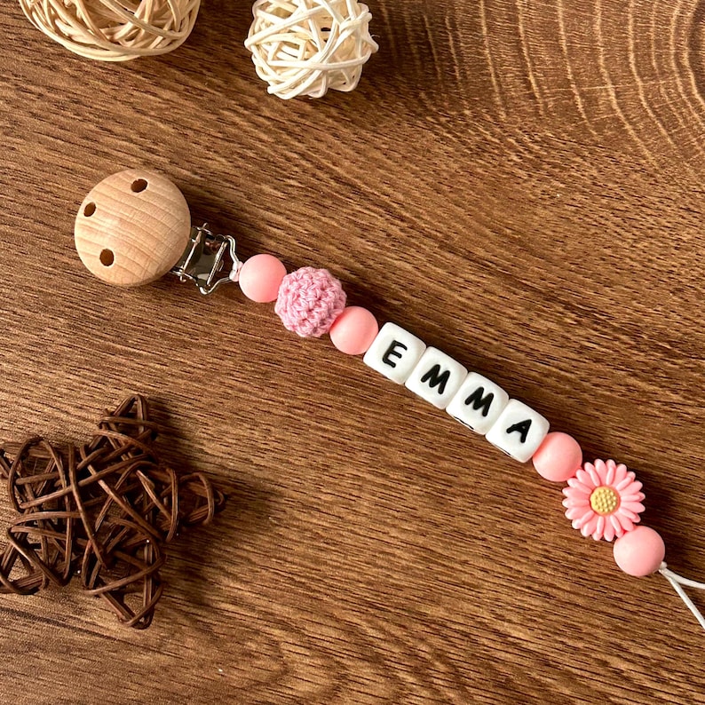 May include: Pink and white baby teether with the name "EMMA" spelled out in white beads. The teether has a wooden clip and a pink crocheted bead.