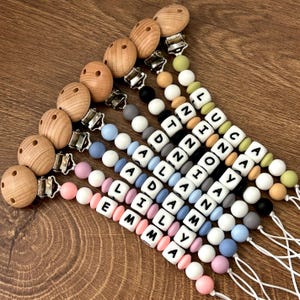 May include: A set of eight personalized silicone baby teethers with wooden clips. Each teether features a different name spelled out in black letters on colorful beads.