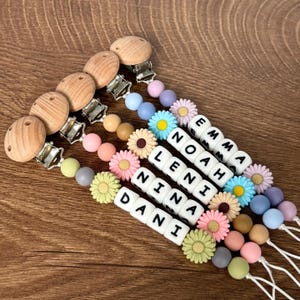 Op de afbeelding: Een verzameling gepersonaliseerde fopspeenclips voor baby's met houten clips en kleurrijke kralen. Elke clip heeft een naam die is gespeld met witte letterkralen en decoratieve bloemkralen. Namen: Dani, Nina, Leni, Noah en Emma.