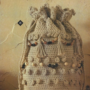 May include: A beige crocheted drawstring bag with wooden and black beads and tassels. The bag is sitting on a wooden surface.