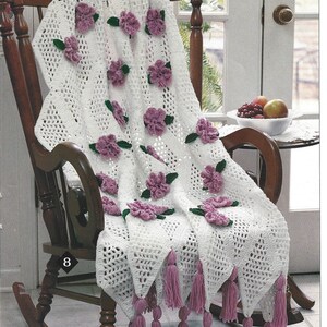 May include: A white crocheted throw blanket with a trellis pattern and purple flowers. The blanket is draped over a wooden rocking chair with pink tassels hanging from the bottom edge. The text "Trellis Roses" is printed below the chair.