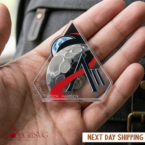 Puede incluir: Un llavero acrílico transparente con un diseño temático espacial. El diseño incluye una luna creciente, la Tierra y una letra "A" estilizada con detalles rojos y negros. Los nombres "Wiseman", "Glover", "Koch" y "Hansen" están impresos.