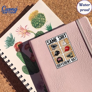 Puede incluir: Un cuaderno rosa de cuero con una pegatina que dice "SAME SHIT DIFFERENT HAT" con im&aacute;genes de diferentes sombreros. Un cuaderno de espiral con ilustraciones de cactus es visible en el fondo. Tambi&eacute;n es visible una pegatina "Water proof".