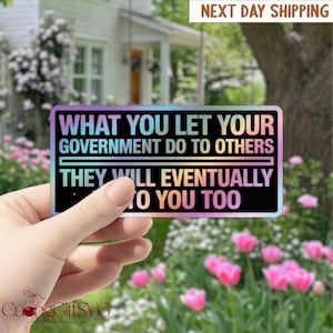Puede incluir: Una pegatina rectangular con el texto "WHAT YOU LET YOUR GOVERNMENT DO TO OTHERS THEY WILL EVENTUALLY TO YOU TOO" en blanco y negro. La pegatina tiene un efecto holográfico, similar al arcoíris. El fondo incluye una casa y flores.