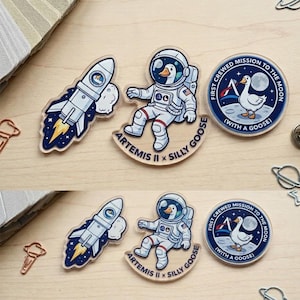 May include: A collection of acrylic pins featuring space-themed designs. One pin depicts a rocket ship, while others showcase an astronaut goose and a circular emblem with a goose on the moon. Text includes "ARTEMIS II x SILLY GOOSE" and "FIRST CREWED MISSION TO THE MOON (WITH A GOOSE)".