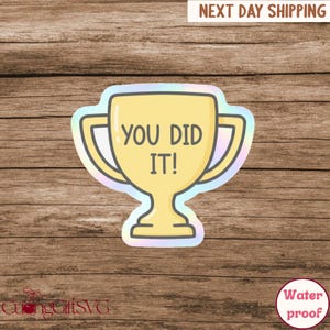 Op de afbeelding: Een gele sticker in de vorm van een trofee met de tekst "YOU DID IT!" in een vetgedrukte lettertype. De sticker heeft een holografische rand en staat op een houten achtergrond. De sticker is waterdicht.