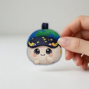 Puede incluir: Una pegatina troquelada que representa un personaje de dibujos animados con un cuerpo de color crema y una cara sonriente. El personaje lleva un sombrero azul con un diseño de mapa del mundo y un gorro de dormir azul marino con estrellas. La pegatina tiene un pequeño lazo en la parte superior.