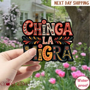 以下が含まれることがあります： カラフルな装飾文字で「CHINGA LA MIGRA」と書かれた黒いステッカー。ステッカーは花の模様で、「Water proof」の文字が見えます。ステッカーは、花と家の背景の前に保持されています。