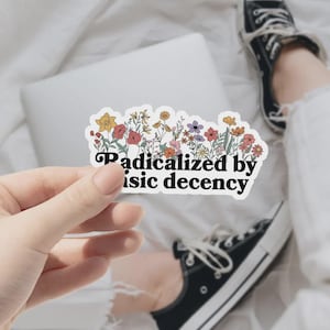 May include: A sticker featuring a floral design and the text "Radicalized by basic decency." The sticker has a variety of colourful flowers, including yellow, red, and purple, arranged above the text. The sticker is held by a hand.