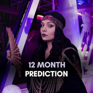 Pode incluir: Uma mulher usando um chapéu preto e uma faixa de cabeça rosa está sentada em uma mesa com uma bola de cristal, cartas de tarô e um leque. O texto "12 MONTH PREDICTION" é exibido em letras brancas sobre um fundo preto.
