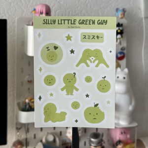 Puede incluir: Una hoja de pegatinas con ilustraciones de dibujos animados de color verde claro. El texto "SILLY LITTLE GREEN GUY" está en la parte superior, con el nombre del artista debajo. La hoja incluye estrellas, diamantes y texto japonés.
