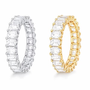 Könnte beinhalten: Zwei Eternity-Ringe, einer silberfarben und einer goldfarben, jeweils mit rechteckigen, klaren Edelsteinen besetzt. Die Ringe werden vor einem weißen Hintergrund präsentiert, der ihre funkelnden Details und ihr elegantes Design hervorhebt.