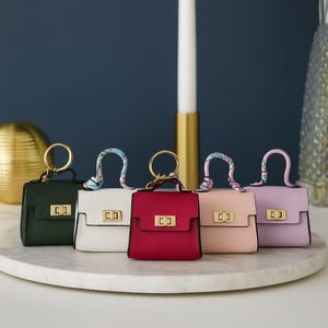 Op de afbeelding: Vijf miniatuurhandtassen in verschillende kleuren: donkergroen, wit, rood, roze en lavendel. Elke tas heeft een goudkleurige sluiting en een bijpassende handgreep met een patroon sjaaltje. De tassen zijn geplaatst op een wit marmeren oppervlak.