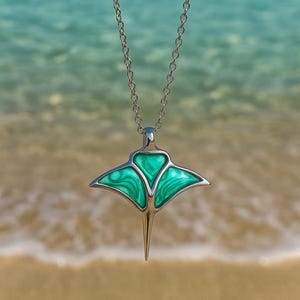 Può includere: Collana color argento con un ciondolo a forma di manta. Il ciondolo presenta un vivace design in smalto verde marmorizzato. Lo sfondo mostra una vista sfocata del mare e della spiaggia.