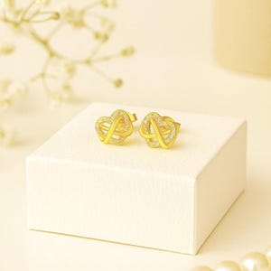 Puede incluir: Un par de pendientes dorados. Cada pendiente presenta un diseño de corazón entrelazado, adornado con pequeñas piedras preciosas transparentes. Los pendientes se exhiben sobre una superficie blanca, con un fondo suave y neutro.