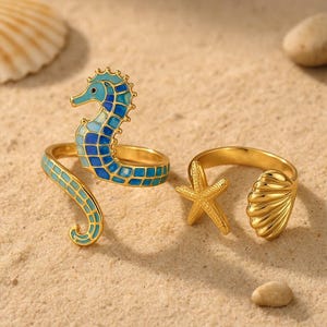 Puede incluir: Anillos dorados con diseños marinos. Uno presenta un caballito de mar con esmalte azul y turquesa, otro una estrella de mar y el tercero una concha. Los anillos se exhiben sobre una superficie arenosa.