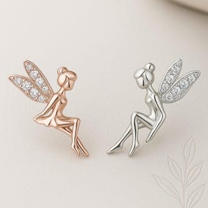 Puede incluir: Dos pendientes con forma de hada. Uno es de oro rosa, el otro plateado. Ambos tienen alas adornadas con cristales transparentes. Las hadas están representadas sentadas con las piernas cruzadas. Los pendientes están sobre un fondo blanco.