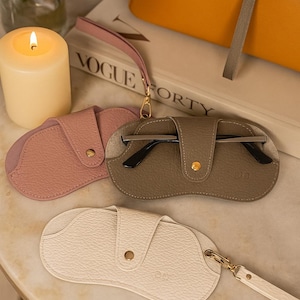 Puede incluir: Tres estuches de gafas de cuero en rosa, topo y crema. Cada estuche tiene una correa de muñeca y un cierre a presión. El estuche topo contiene un par de gafas con montura negra. Una vela encendida y un bolso amarillo están en el fondo.