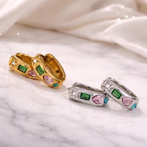May include: Two pairs of hoop earrings. One pair is gold-toned, the other is silver-toned. Each earring features a rectangular green, pink, and blue gemstone. The earrings are on a white marble surface.