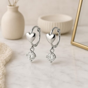 Puede incluir: Un par de pendientes de aro plateados. Cada pendiente presenta un dije en forma de corazón en la parte superior y una gema cuadrada y brillante colgando debajo. Los pendientes se exhiben sobre una superficie de mármol.