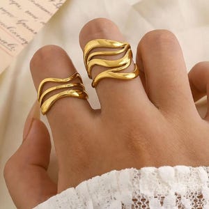 Puede incluir: Dos anillos dorados con un diseño ondulado se muestran en dedos. Los anillos tienen una apariencia en capas, con múltiples bandas que crean una estética texturizada y fluida. Los anillos se colocan sobre un fondo suave y neutro.