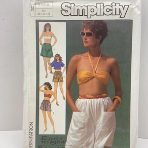 Puede incluir: Un paquete de patrones de costura Simplicity vintage, que muestra a una mujer con un top halter amarillo y pantalones cortos blancos. El paquete incluye ilustraciones de diferentes combinaciones de pantalones cortos y tops. El texto "E-A-S-Y TO SEW" es visible.