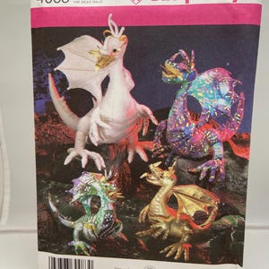 May include: Simplicity pattern envelope showing four dragon figures in various colours. The dragons are white, gold, green, and purple, with detailed wings and scales. The envelope has the number 4063 and the text "ONE SIZE".
