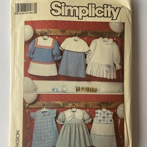 May include: A Simplicity sewing pattern envelope, number 7983, size 4, featuring illustrations of six different children's dresses. The designs include various blue and white fabrics with collars, ruffles, and patterns like stripes and hearts.