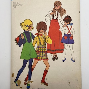 Op de afbeelding: Vintage Simplicity naaipatroon, maat 10, voor meisjes. De omslag toont illustraties van vier verschillende outfits, waaronder een rode jurk, een groene overall en een gele geruite outfit. De tekst op de omslag luidt "Simplicity 9542" en "Taille 62.2 cm".