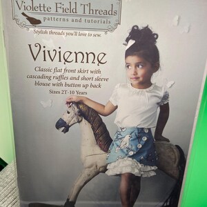 May include: A sewing pattern package from Violette Field Threads features a girl wearing a skirt and blouse. The package is labeled "Vivienne" and includes instructions for a classic flat front skirt with cascading ruffles and a short sleeve blouse. Sizes 2T-10 Years.
