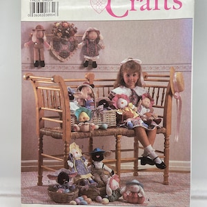 Puede incluir: Patrón de Simplicity Crafts con una variedad de muñecas hechas a mano y artículos decorativos. La portada muestra a una niña rodeada de muñecas, cestas y un banco de madera. El patrón incluye instrucciones para crear estas manualidades.