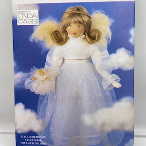 May include: Vogue Craft pattern for an angel doll. The doll is wearing a white dress with a sheer overlay, and has blonde hair and blue eyes. The pattern includes a full-color iron-on transfer for the face. The packaging is blue with white clouds.