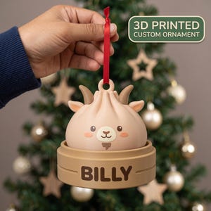 Könnte beinhalten: Ein 3D-gedrucktes, personalisiertes Ornament in Form einer hellbeigen Ziegenfigur mit kleinen Hörnern, Ohren und einem Ziegenbart, die in einem hellbraunen Bambusdampfer-Sockel mit dem aufgedruckten Namen BILLY sitzt und an einem roten Band hängt.