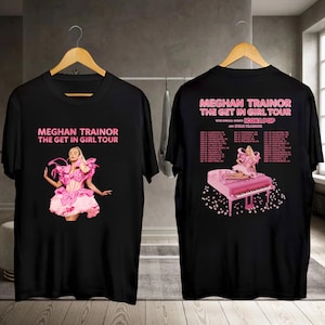 May include: Black t-shirts featuring the text "MEGHAN TRAINOR THE GET IN GIRL TOUR" in pink. One shirt displays a pink-clad figure, the other lists tour dates and a pink piano graphic. The shirts are hanging on a wooden hanger.