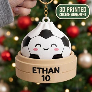 Puede incluir: Un adorno personalizado impreso en 3D con forma de balón de fútbol blanco y negro diseñado como una empanadilla con cara sonriente y mejillas rosadas, sobre una base de vaporera color canela con el texto ETHAN 10 impreso en negro al frente.
