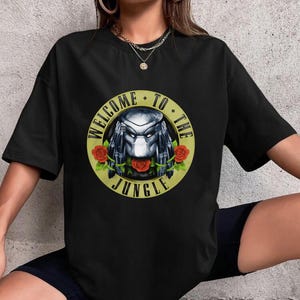 May include: Black t-shirt featuring a circular design with the text "WELCOME TO THE JUNGLE". The design includes a Predator-like character with roses. The shirt is a crew neck and has short sleeves.