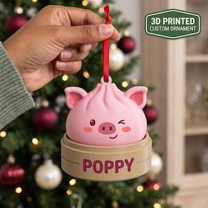 Könnte beinhalten: Ein 3D-gedrucktes, personalisiertes Ornament in Form eines rosa Schweine-Dumplings, das in einer beigen Bambusdampfer-Basis sitzt. Die Basis trägt den Namen POPPY in dunkelroten Buchstaben. Ein rotes Band dient zum Aufhängen am Weihnachtsbaum.