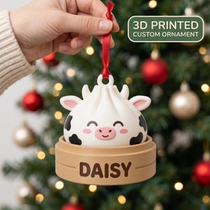 Puede incluir: Un adorno personalizado impreso en 3D que presenta un personaje de bola de masa blanco con manchas de vaca, mejillas rosadas y una cara sonriente, sentado en una base de vaporera de bambú color canela con el nombre DAISY, colgado de una cinta roja.