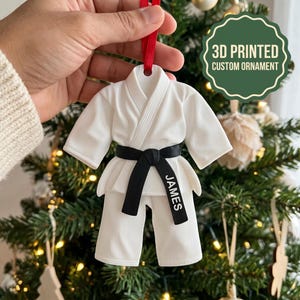 Op de afbeelding: Een wit 3D-geprint ornament in de vorm van een vechtsportuniform met een zwarte band waarop de naam JAMES in witte letters staat. Het ornament hangt aan een rood lint en toont de tekst 3D PRINTED CUSTOM ORNAMENT in de rechterbovenhoek.