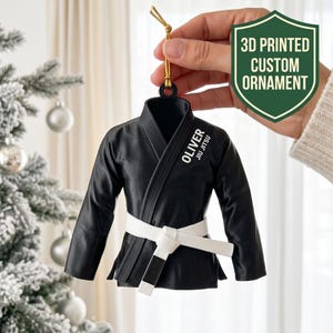 Puede incluir: Un adorno de kimono de artes marciales negro impreso en 3D con un cinturón blanco y el texto OLIVER JIU JITSU impreso en blanco en el pecho. El adorno cuelga de un cordón dorado e incluye un pequeño lazo en la parte superior para colgarlo.