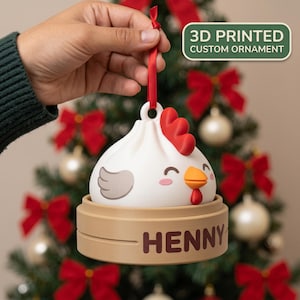 Puede incluir: Un adorno personalizado impreso en 3D con forma de bola de masa de pollo blanca con cresta roja, pico naranja y ala gris. La base es una vaporera de bambú color canela con el nombre HENNY en letras marrón oscuro, suspendido por una cinta roja.